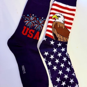 May include: Two navy blue socks. One sock features the word "USA" in red with fireworks. The other sock has a design of the American flag with a bald eagle. The socks are a patriotic theme.