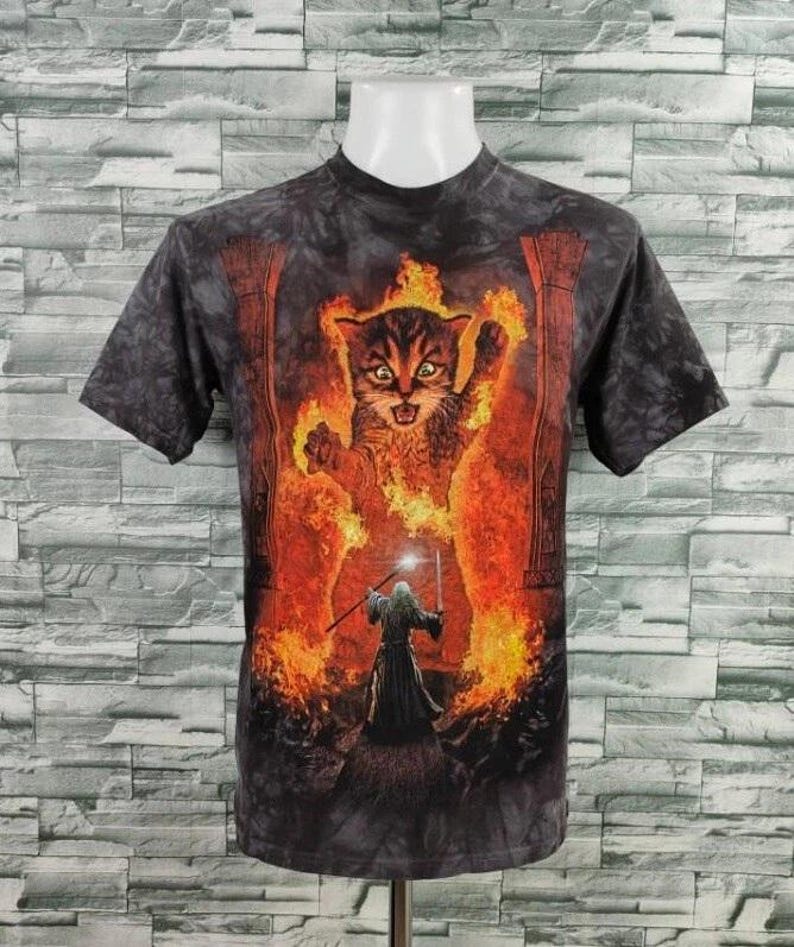 May include: A short-sleeved, dark gray t-shirt featuring a fantasy scene. The graphic depicts a fiery cat with a wizard holding a staff. The cat is surrounded by flames, and the wizard stands in front of it.