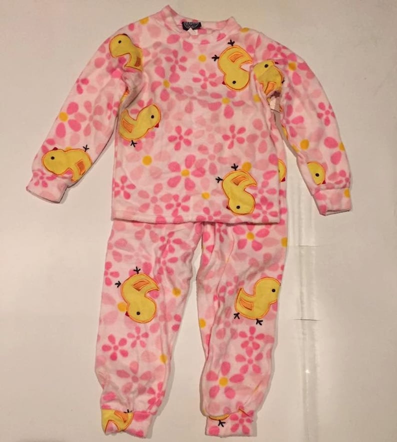 May include: Pink and yellow pajamas with a floral pattern and yellow ducklings. The pajamas are long-sleeved and have a crew neck.