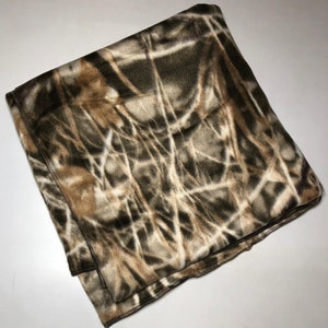 Puede incluir: Una manta de forro polar de camuflaje marrón, beige y blanco. La manta está doblada por la mitad y tiene una textura suave y peluda.