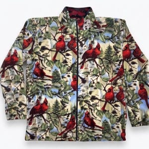 May include: A long-sleeve zip-up fleece jacket featuring a repeating pattern of bright red cardinal birds perched on tree branches against a backdrop of white birch trees and green foliage. The jacket has a high collar and a black front zipper.