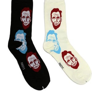 Op de afbeelding: Twee paar novelty sokken, één zwart en één crème, elk met een herhaald patroon van Abraham Lincoln's gezicht in rood en blauw. Het woord "FOOZYS" staat op de zijkant van elke sok.
