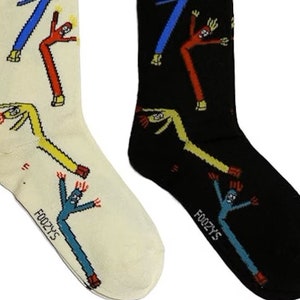 May include: A pair of socks with a fun design featuring inflatable tube men in various colors. One sock is white with yellow, red, and blue tube men. The other sock is black with yellow, red, and blue tube men. The socks have the word "Foozys" printed on the bottom.