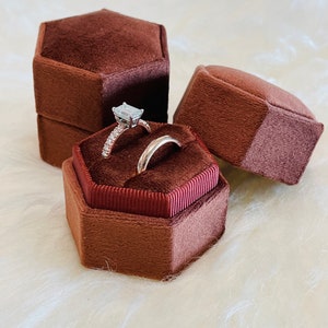 Puede incluir: Dos anillos en una caja de anillo hexagonal de terciopelo marrón. El anillo de compromiso tiene un diamante de forma cuadrada y una banda con pequeños diamantes. La banda de boda es una banda lisa.