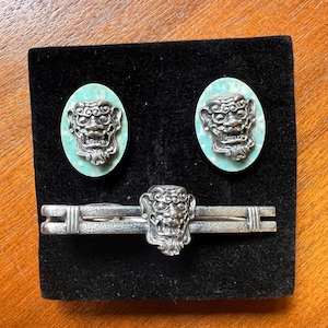 May include: A set of jewelry featuring a silver-toned tie clip and matching oval earrings. Each piece has a turquoise-colored background with a detailed, silver-toned face design. The set is displayed on a black velvet surface.