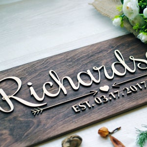 Personalized wooden family name sign featuring 'Richardson' in raised 3D script lettering. A decorative arrow and heart design separates the name from the date 'EST. 03.17.2017' below. The sign features a dark stained wood background with light, natural wood letters.
