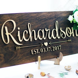 Rustic wooden sign reading 'Richardson' in raised cursive letters with a heart and arrow design and established date of 2017. The sign is stained dark walnut and displayed on a matching wooden stand next to white flowers.