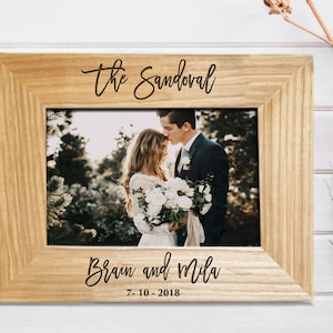 Personalized wooden photo frame engraved with the family name 'The Sandoval' in a flowing script font at the top. The bottom features the couple's first names, 'Brian and Mila,' and the date '7-10-2018.' The frame holds a wedding portrait of a groom kissing a bride on the forehead, set against a background of white wood and dried cotton flowers.