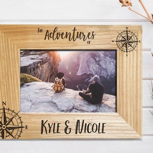 May include: A wooden photo frame with a compass design and the text "The Adventures of Kyle & Nicole". The frame has a photo of a couple standing on a mountaintop.
