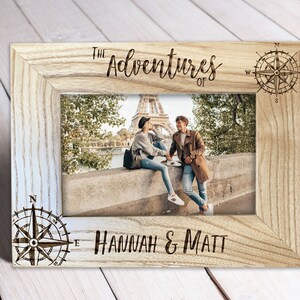 May include: Wooden photo frame with the text "The Adventures of Hannah & Matt" and compass rose designs. The frame holds a photo of a couple in front of the Eiffel Tower. The frame is made of light-colored wood.
