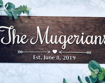 May include: A dark brown wooden sign with the text "The Mugerians Est. June 8, 2019" in white lettering. The text is in a cursive font and is centered on the sign. There are two arrows pointing towards a small heart in the middle of the sign.