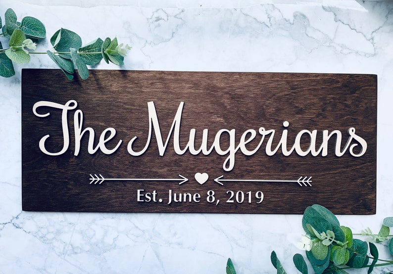Personalized wooden family name sign featuring "The Mugerians" in raised 3D white script lettering on a dark walnut stained background. Below the name is a decorative arrow and heart graphic, followed by "Est. June 8, 2019" in serif font.