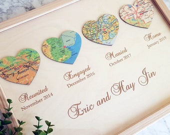 May include: A wooden frame with a light brown background. Five heart-shaped maps are displayed on the frame, each representing a different milestone in a couple's relationship. The text "Reunited November 2014", "Engaged December 2016", "Married October 2017", "Home January 2019", and "Eric and Hay Jin" is inscribed on the frame.