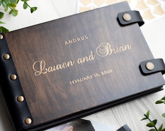 May include: A dark brown wooden photo album with black leather trim and gold-colored metal accents. The album is engraved with the text "Lauren and Brian" and "February 16, 2020" in gold lettering.