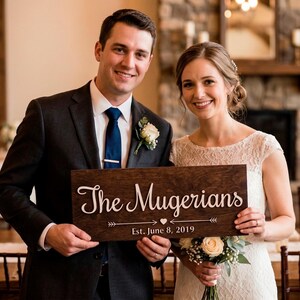 A smiling bride and groom holding a personalized rectangular dark wood sign. The sign features the white laser-cut text "The Mugerians" in a cursive script, with "Est. June 8, 2019" and a decorative arrow design below it. The couple is in a rustic indoor venue with a stone fireplace and candlelit tables in the background.