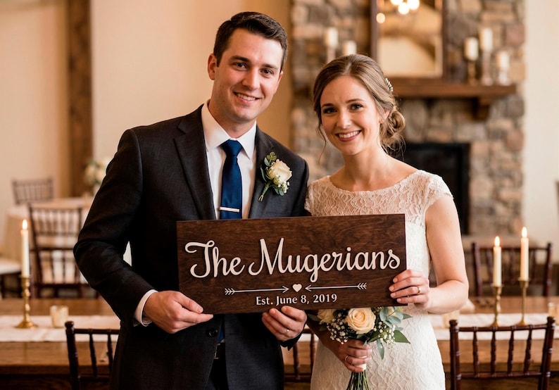 A smiling bride and groom holding a personalized rectangular dark wood sign. The sign features the white laser-cut text "The Mugerians" in a cursive script, with "Est. June 8, 2019" and a decorative arrow design below it. The couple is in a rustic indoor venue with a stone fireplace and candlelit tables in the background.