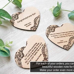 Three personalized wooden heart-shaped gift notes featuring laser-engraved messages and floral lace corners, arranged on a white wood background with eucalyptus leaves. A text overlay at the bottom reads: "For each of your orders, you can add this beautiful wooden note for free! Make your order even more unique.