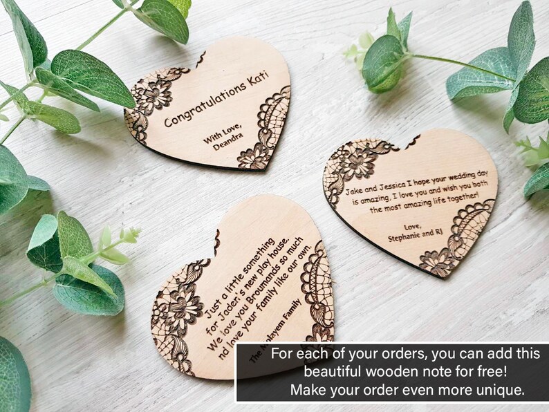 Three personalized wooden heart-shaped gift notes featuring laser-engraved messages and floral lace corners, arranged on a white wood background with eucalyptus leaves. A text overlay at the bottom reads: "For each of your orders, you can add this beautiful wooden note for free! Make your order even more unique.