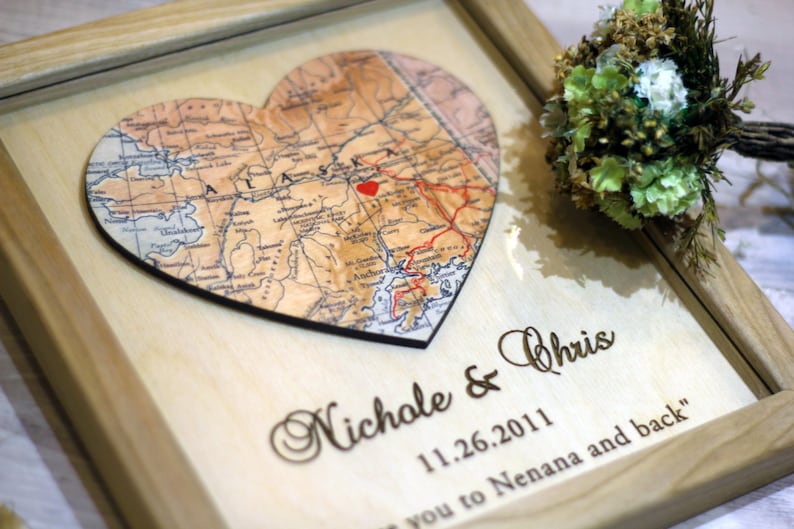 Personalized wooden picture frame featuring a laser-engraved heart-shaped map of Alaska with a red heart marking a special location. Custom engraved with couple's names 'Nichole & Chris' and wedding date '11.26.2011' below the map. Rustic wood frame with a 'love you to Nenana and back' quote, perfect for a custom wedding or anniversary gift.