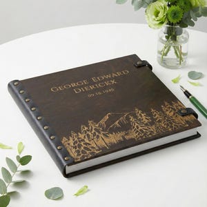 A dark wood personalized photo album with a black leather spine. The cover is engraved with the name George Edward Dierickx, a 1945 date, and a beautiful mountain and pine forest landscape, lying next to a green pen.