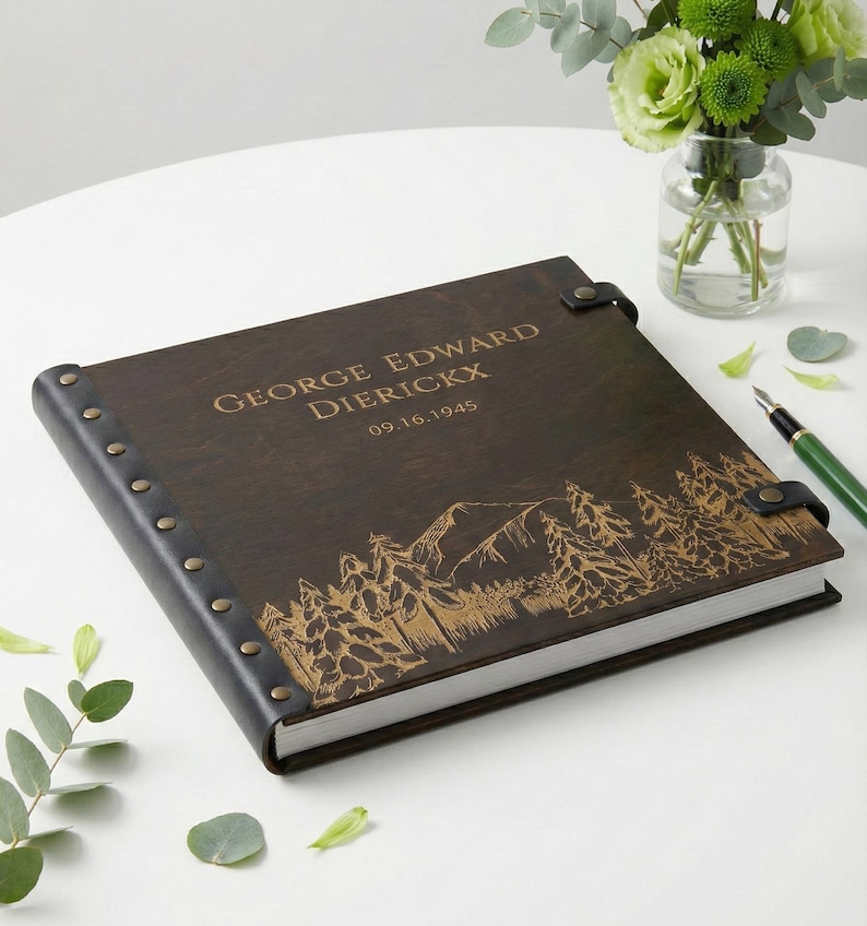 A dark wood personalized photo album with a black leather spine. The cover is engraved with the name George Edward Dierickx, a 1945 date, and a beautiful mountain and pine forest landscape, lying next to a green pen.