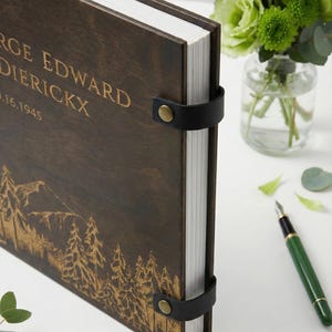 Rustic wood and leather photo album with a detailed forest and mountain engraving. The dark wood cover is personalized with engraved lettering and bound with black leather and brass rivets.