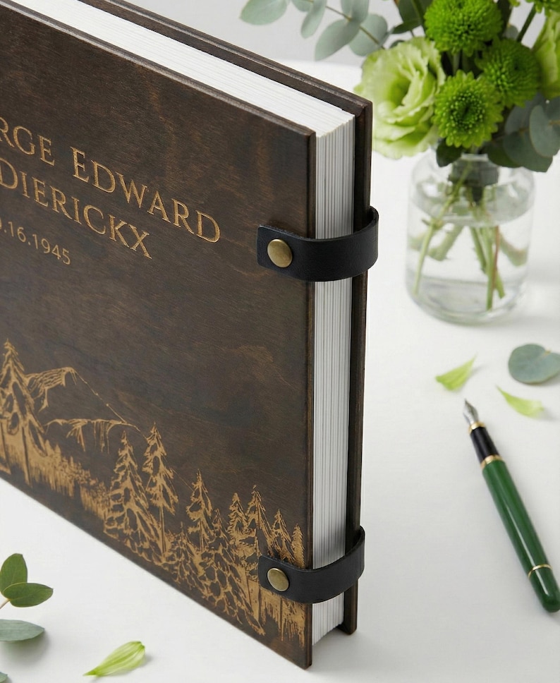 Rustic wood and leather photo album with a detailed forest and mountain engraving. The dark wood cover is personalized with engraved lettering and bound with black leather and brass rivets.