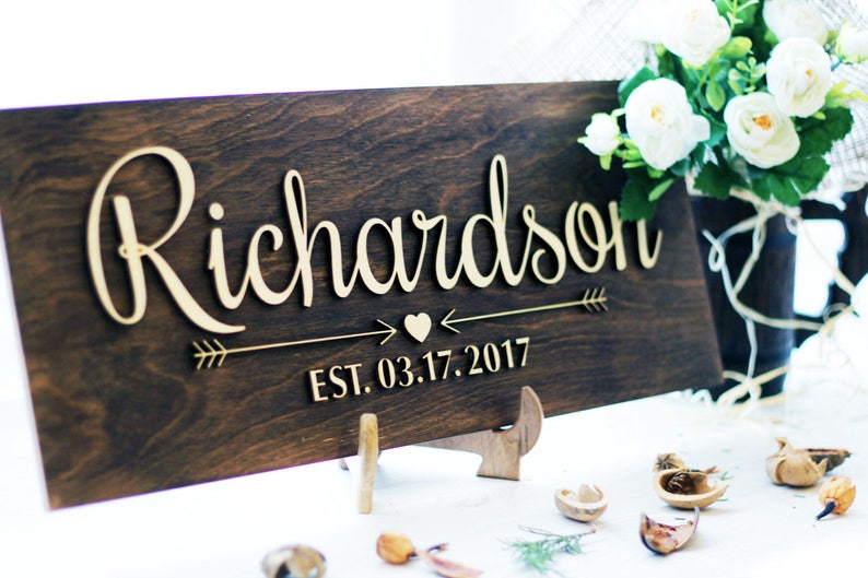 Rustic wooden sign reading 'Richardson' in raised cursive letters with a heart and arrow design and established date of 2017. The sign is stained dark walnut and displayed on a matching wooden stand next to white flowers.