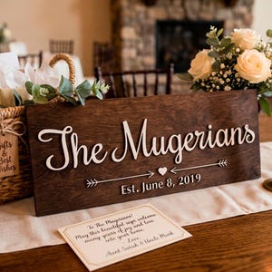 A dark-stained rectangular wooden sign with raised white 3D lettering that reads "The Mugerians" in a cursive font. Below the name is a decorative arrow with a heart and "Est. June 8, 2019." The sign is part of a rustic wedding or anniversary table display, featuring a woven gift basket, white roses, lit taper candles in brass holders, and a personalized greeting card. This handcrafted piece highlights the laser-engraved details and elegant design.