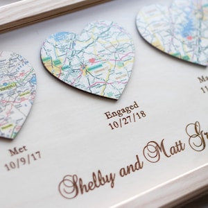 May include: A white wooden frame with three heart-shaped maps inside. The maps are of different locations and are labeled with the dates "10/9/17", "10/27/18", and "11/23/19". The text "Met", "Engaged", and "Married" are also included. The names "Shelby and Matt Grant" are written in cursive at the bottom of the frame.