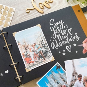 A hand writing with a silver metallic pen in a black-paged wedding Photo album. The open album features a large white engraved moon and the quote 'I love you to the moon and back.' Several wedding photos are mounted with gold corner stickers. A wooden 'love' sign and photo corners sit on the light wood table in the background.