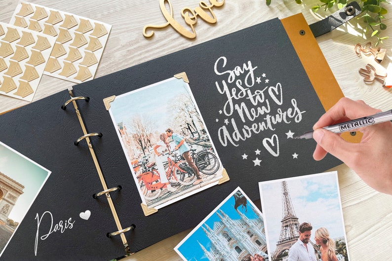 A hand writing with a silver metallic pen in a black-paged wedding Photo album. The open album features a large white engraved moon and the quote 'I love you to the moon and back.' Several wedding photos are mounted with gold corner stickers. A wooden 'love' sign and photo corners sit on the light wood table in the background.