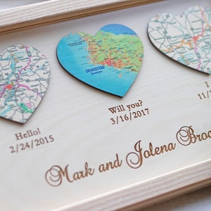May include: Wooden framed artwork featuring three heart-shaped map cutouts. The artwork includes the text "Hello! 2/24/2015", "Will you? 3/16/2017", and "I do! 11/23/2019". The names "Mark and Jolena Brocious" are also engraved on the wood.