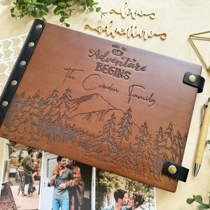 Personalized brown leather photo album with custom engraving. The cover features a laser-engraved mountain and forest landscape with the text "And So The Adventure Begins - The Cowden Family." It includes a black leather spine with brass rivets, matching leather closures, and sits next to a wooden pen and travel photos.