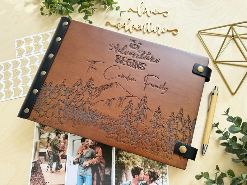 Personalized brown leather photo album with custom engraving. The cover features a laser-engraved mountain and forest landscape with the text "And So The Adventure Begins - The Cowden Family." It includes a black leather spine with brass rivets, matching leather closures, and sits next to a wooden pen and travel photos.
