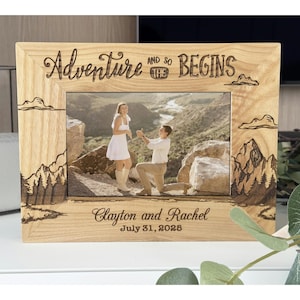 May include: Wooden picture frame with the words "Adventure and so the BEGINS" engraved at the top. The frame holds a photo of a couple, with the names "Clayton and Rachel" and the date "July 31, 2025" engraved at the bottom. The frame has mountain and cloud designs.