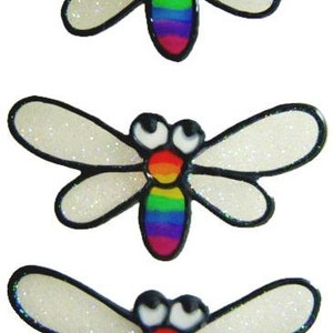 以下が含まれることがあります： 虹色の羽根と黒い輪郭を持つ、白いグリッターのトンボのステッカーが3つ。トンボは大きく黒い目と赤い口をしています。