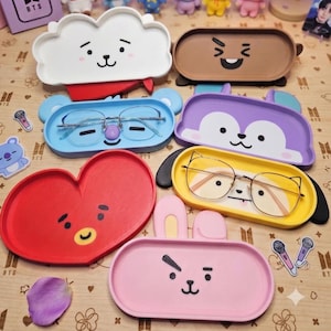 Puede incluir: Una colección de siete bandejas coloridas con temas de personajes, que presentan varias formas y expresiones faciales. Las bandejas incluyen una nube blanca, un rectángulo marrón, un óvalo azul, una forma morada, un corazón rojo, un óvalo amarillo y un óvalo rosa, con dos pares de gafas de montura metálica descansando dentro de las bandejas azul y amarilla.