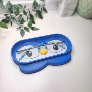 May include: A blue eyeglasses holder shaped like a cartoon penguin, with a white interior and a yellow beak. The holder contains a pair of eyeglasses with a silver frame and light blue temples. The holder is on a white surface.