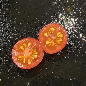 May include: Two halves of a ripe, red cherry tomato, showcasing the juicy interior and seeds. The cut surfaces reveal the tomato's structure, with a star-like pattern. The background is a dark, reflective surface with water droplets.