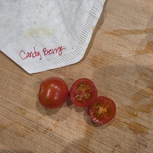 May include: A whole red cherry tomato and two halves on a wooden cutting board. A white cloth with the words "Candy Berry" in red script is in the upper left corner. The tomatoes are ripe and juicy.