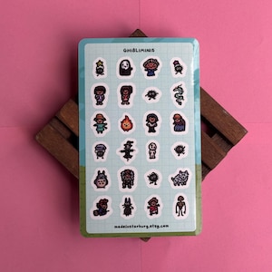 May include: A sticker sheet with cartoon characters, including a faceless spirit, soot sprites, and forest spirits. The stickers are on a light blue background with the text "GHIBLIMINIS" and "madeinstarburg.etsy.com". The sheet is on a wooden stand against a pink background.