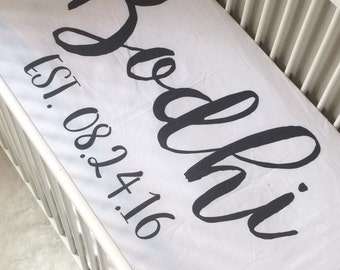 May include: White crib sheet with black cursive text that reads "Bodhi" and "Est. 08.24.16".