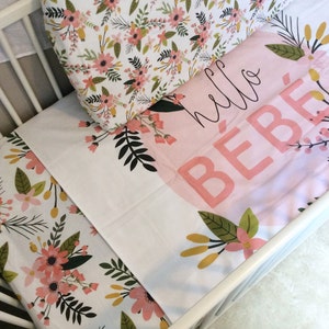 May include: A white cot with a floral patterned fitted sheet and a pink and white blanket with the text "hello bebe" on it.