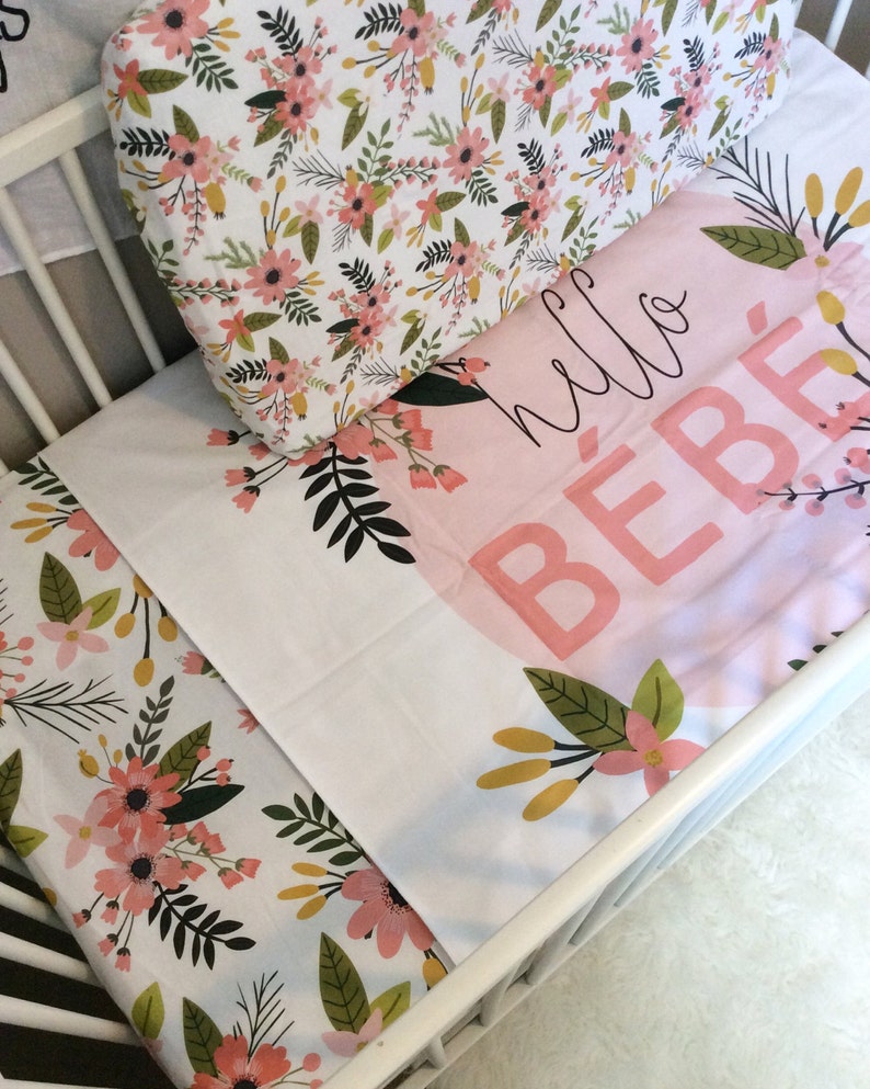 May include: A white cot with a floral patterned fitted sheet and a pink and white blanket with the text "hello bebe" on it.