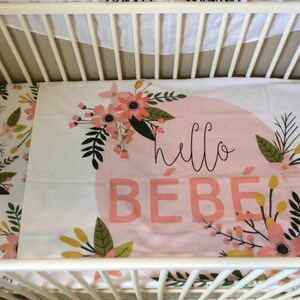 May include: A white cot with a pink and white floral cot sheet. The sheet has a pink circle with the text "hello b&eacute;b&eacute;" in black.