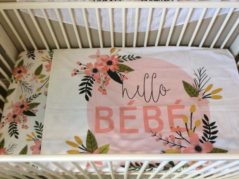 May include: A white cot with a pink and white floral cot sheet. The sheet has a pink circle with the text "hello b&eacute;b&eacute;" in black.
