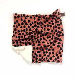 May include: A soft, plush blanket with a coral pink background and black leopard print. One side of the blanket is made of a fluffy white material.