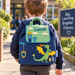 May include: A navy blue quilted backpack featuring a green flap with the name Lucas, a castle graphic, and clouds. The main body displays a green dragon with yellow wings breathing fire, two small trees, and a shield-shaped charm.