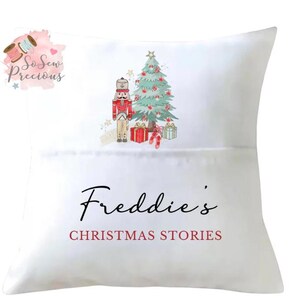 Könnte beinhalten: Weißes Kissen mit Weihnachtsmotiv. Es zeigt eine Aquarellillustration eines Nussknackers, eines Weihnachtsbaums und Geschenken. Der Text "Freddie's CHRISTMAS STORIES" ist in Schwarz und Rot gedruckt.
